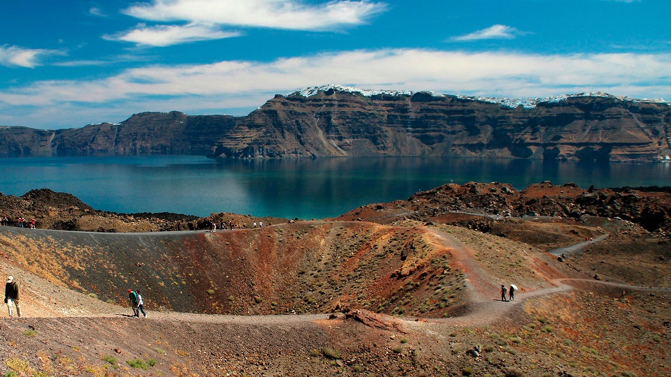 Paisaje donde se observa hacia atrás un cerro, luego un lago y hacia adelante un camino con personas caminando.