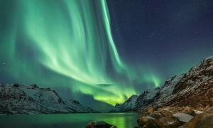 Aurora boreal de tonos verdosos y amarillos en el cielo. Alrededor montañas y un lago que refleja la aurora del cielo. Tomada en islandia