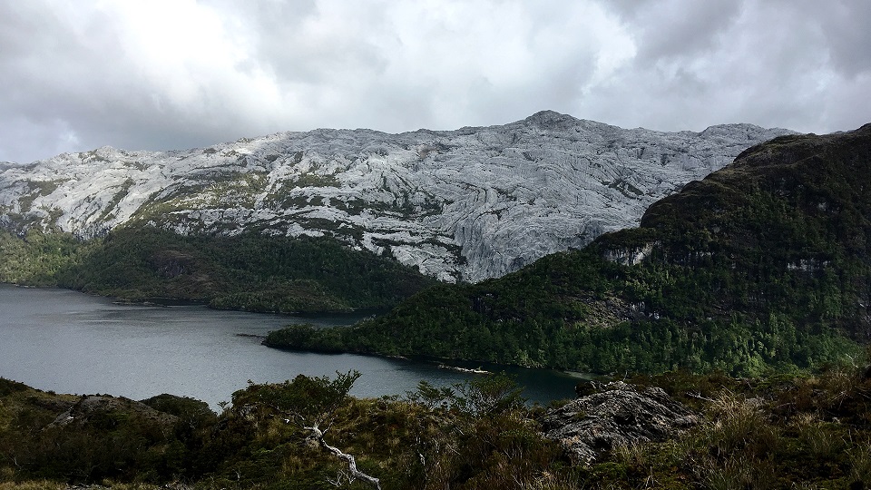 Fotografía: Vista de un cerro donde se pueden ver plieguesm abajo un hermoso lago y alrededor vegetación frondosa. Foto del sur de Chile